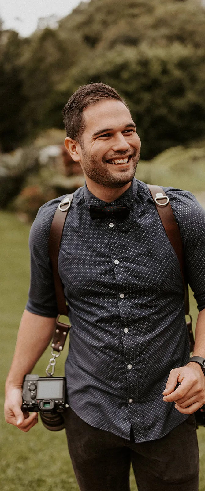Sean Harman smiling, wearing a bow tie with two cameras on a leather harness in a garden